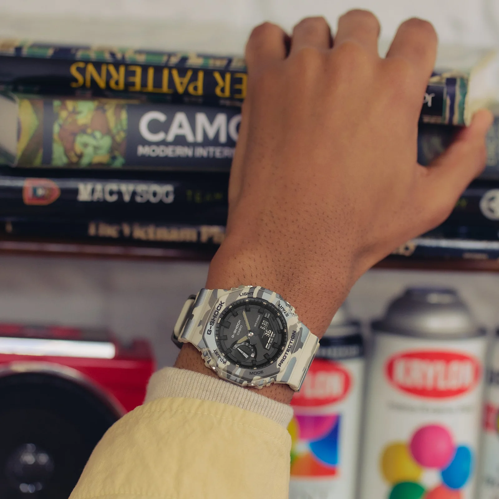 Ein Mann mit einer gelben Jacke trägt die GA-2100CM-5AER am Handgelenk und greift in ein Bücherregal