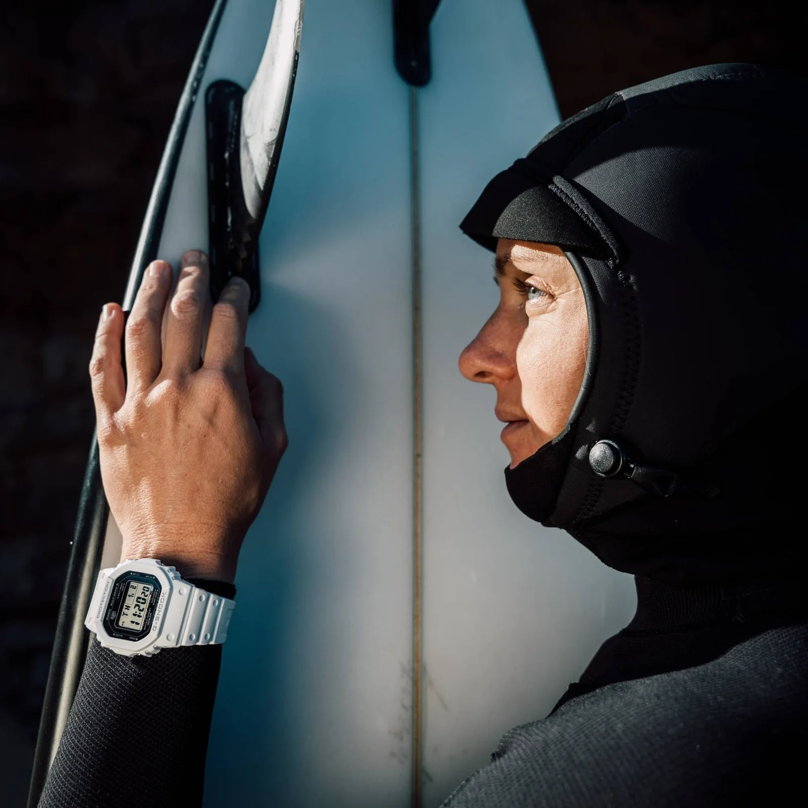 Eine Frau mit Neoprenanzug steht neben einem Surfbrett und trägt die weiße GW-5000HS-7ER mit schwarzem Zifferblatt am Handgelenk