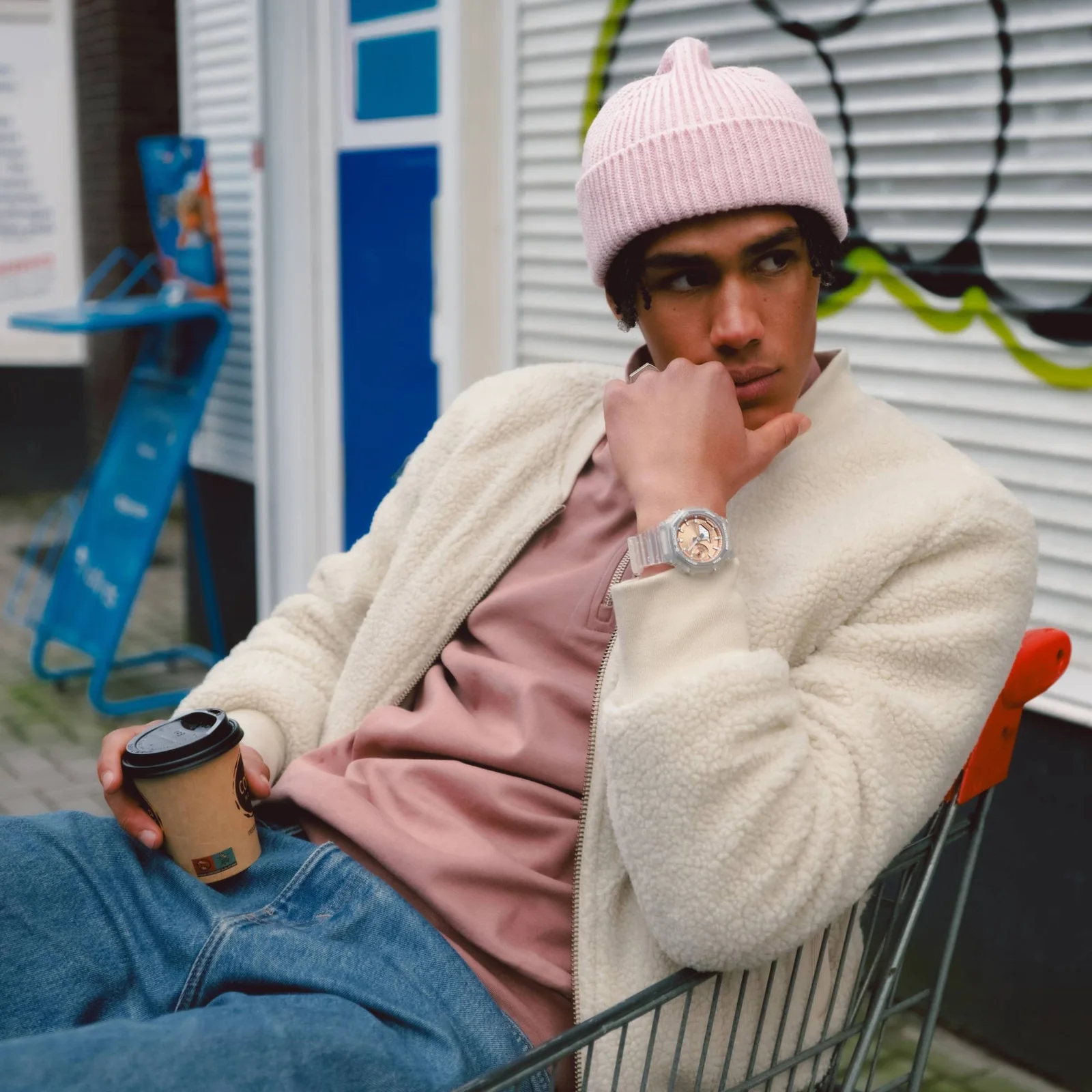 Das männliche Model sitzt in einem Einkaufswagen und trägt die die GA-2100BM-7A5 in Kombination mit einem modischen Streetwear-Outfit bestehend aus pastellfarbenen Beanie und T-Shirt sowie einer weißen Jacke.