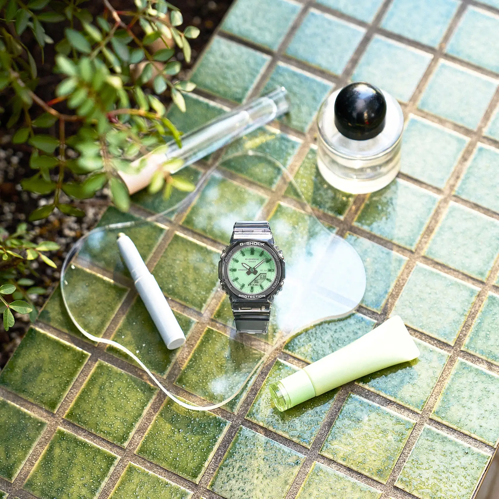 Die G-SHOCK Armbanduhr GMA-P2100SR-1AER liegt auf einer grün gefliesten Fläche mit Kosmetikartikeln daneben
