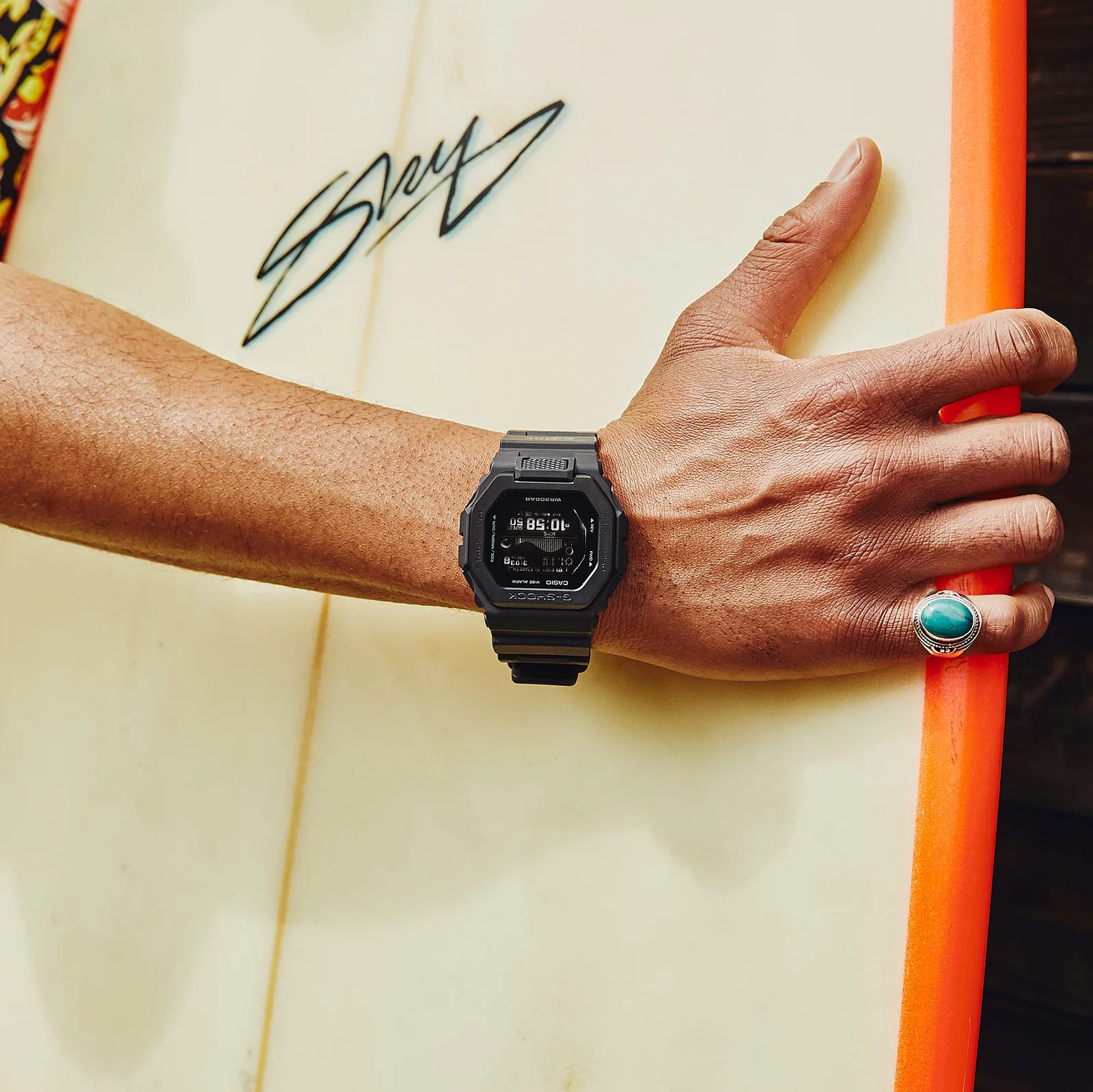 Die GBX-100NS-1ER am Handgelenk eines Mannes, der ein Surfbrett in der Hand hält
