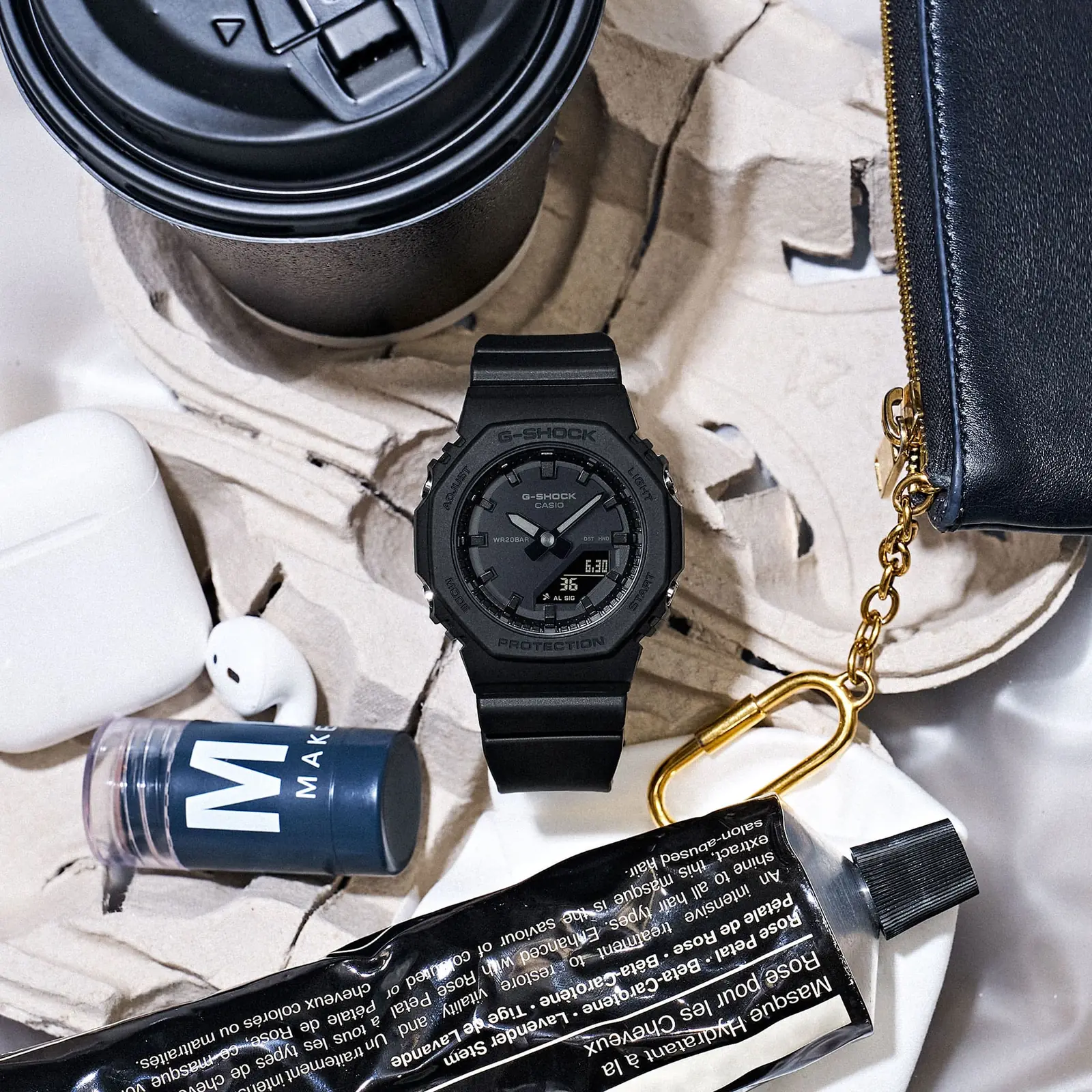 Die schwarze Armbanduhr GMA-P2100BB-1A liegt neben einem Coffee-to-Go Becher und verschiedenen Handtaschen-Utensilien