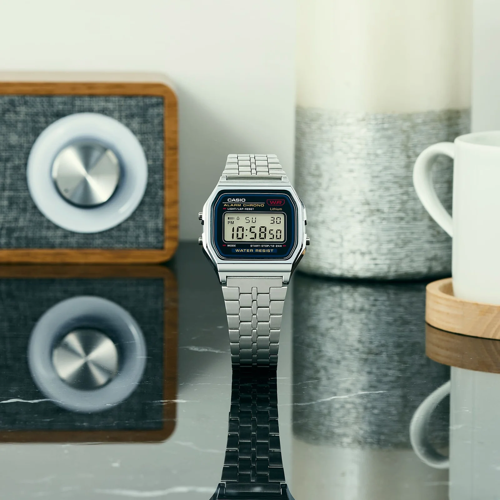Die digitale Armbanduhr A159WE-1EF steht auf einer spiegelnden Tischoberfläche mit einem modernen Radio und einer Tasse im Hintergrund