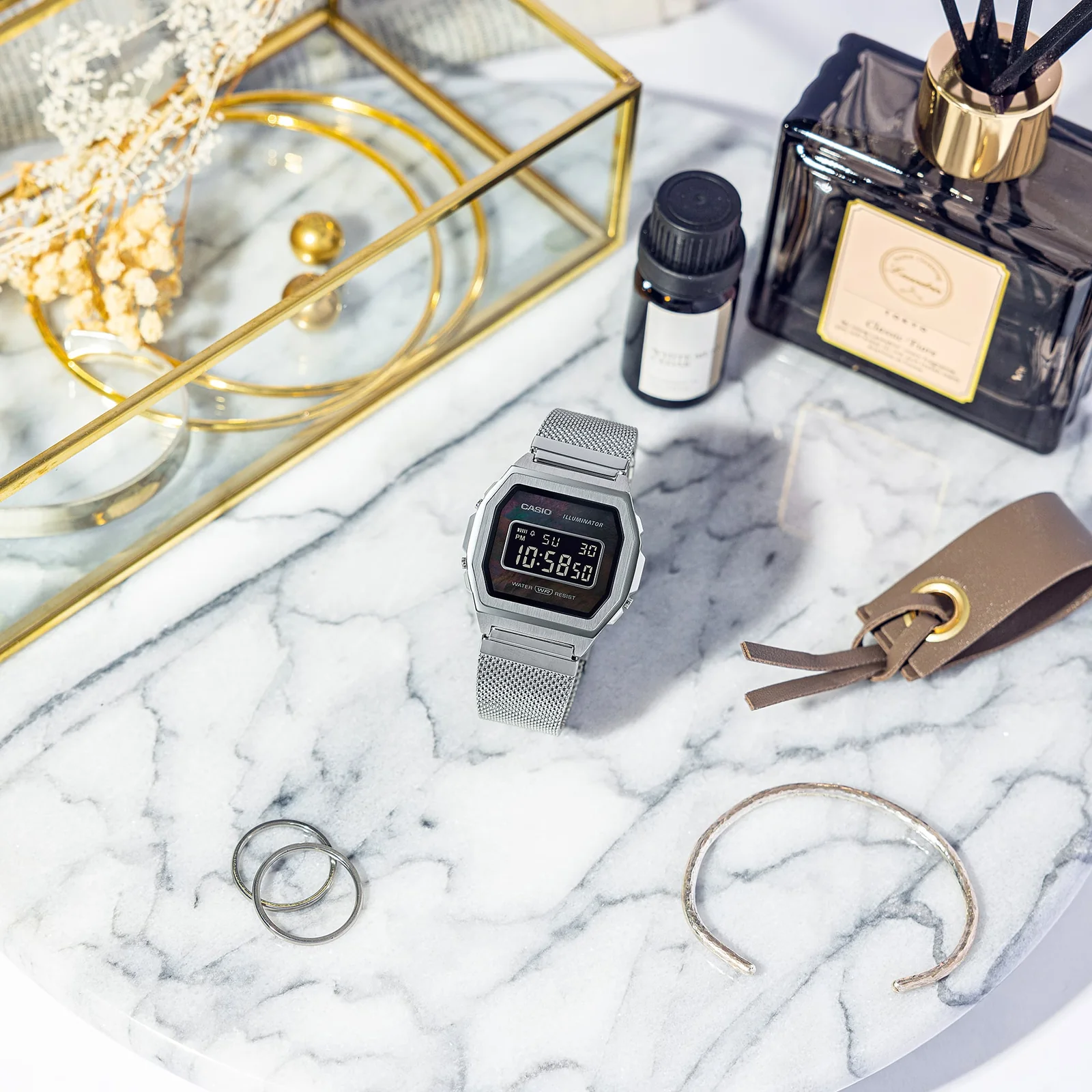 A1000M-1B Digitaluhr mit Mesh-Edelstahlband auf Marmorplatte umgeben von Schmuck, Raumduft und Accessoires