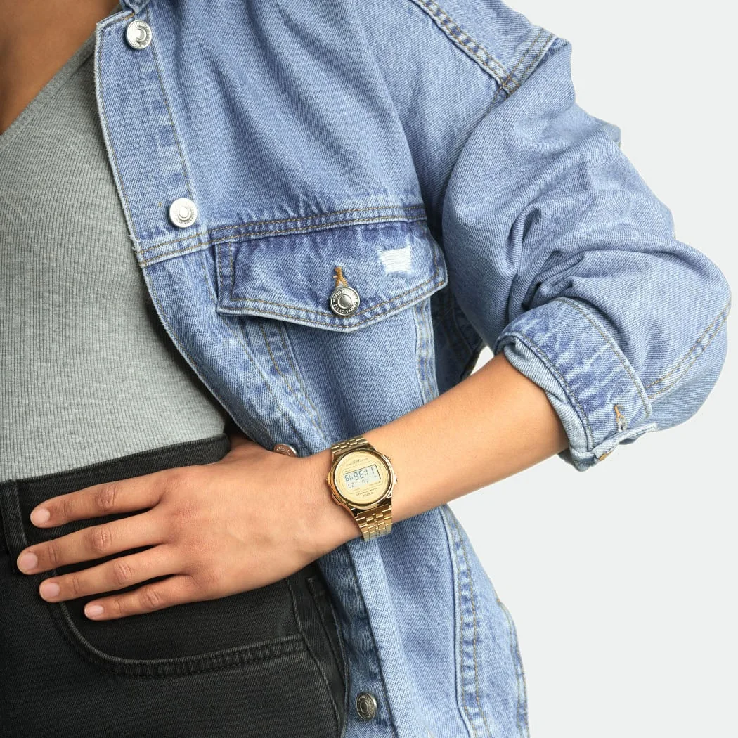 Frau trägt eine runde, goldene Mesh-Armbanduhr, kombiniert mit einer Jeansjacke und einem grauen Oberteil